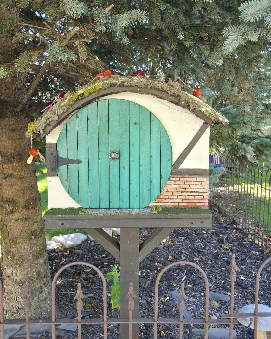 A handmade book-sharing box under a blue spruce: a round green plank door with a black strap hinge, cream walls, a brick corner and a curved mossy roof, on a timber post.